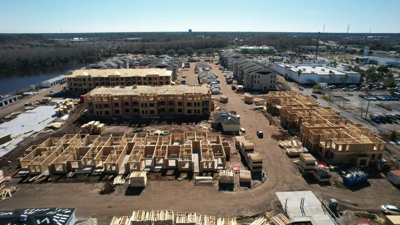 Aerial view across a construction site showing rows of first-floor wall framing in the foreground and a three-story building with its Howland roof trusses already set behind them.