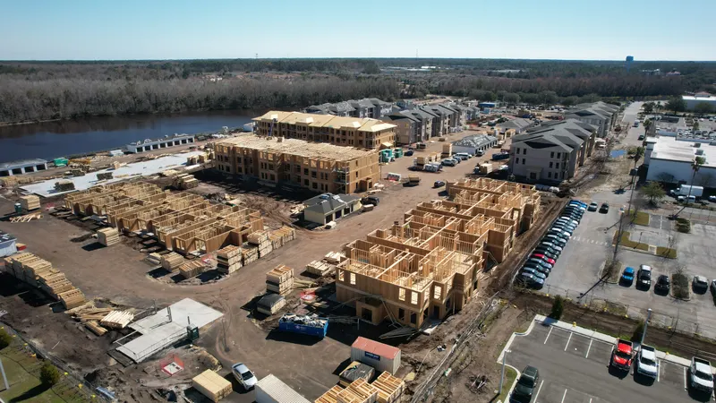 Elevated aerial of a residential construction site beside a river, with partially framed buildings in the center and finished dark-roofed buildings behind.