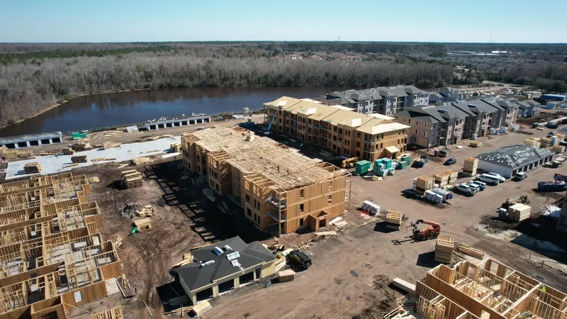 Aerial view of a three-story building with Howland roof trusses set and roof sheathing underway, with completed buildings and a pond behind it.