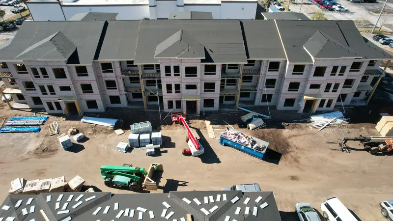 Aerial view of a three-story building dried in with roof underlayment and house wrap, with a boom lift and debris container in front of it.