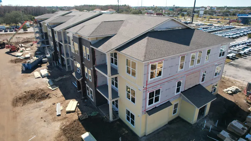 Aerial view of a three-story building with a shingled hip roof, house wrap, and the first sections of yellow lap siding installed.