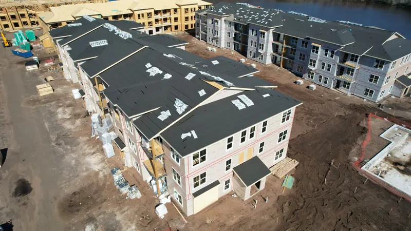 Aerial view of two three-story buildings with black roof underlayment and sheathed walls, water visible along the back of the site.