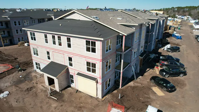 Aerial corner view of a three-story building with its roof shingled and walls wrapped, taped window openings visible and trucks parked alongside.