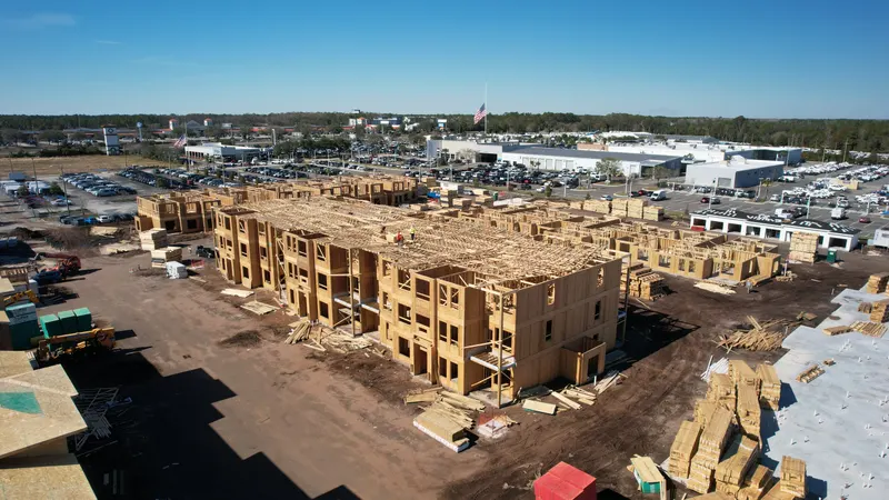 Aerial view of a large building block in bare wood framing with Howland roof trusses set, and truss and lumber bundles staged in the adjacent yard.