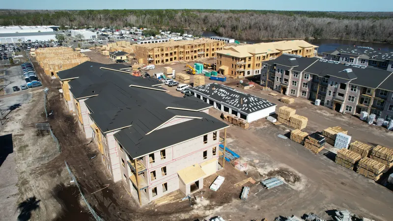 Aerial view across the site with a fully roofed building in the foreground and stacks of Howland truss and lumber bundles staged to one side.