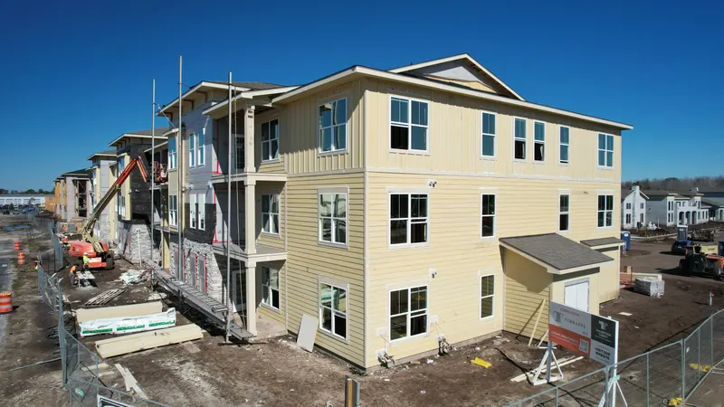 Low aerial of a three-story building with yellow lap siding installed, scaffolding at the far end and construction fencing around it.