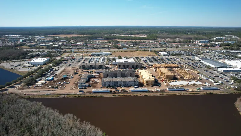 High aerial of the whole construction site beside a wide river, with buildings ranging from bare framing to finished roofs.