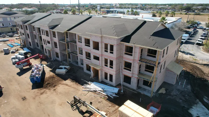 Aerial view of a long three-story building with black roof underlayment and wrapped walls, a lift and debris container in front.